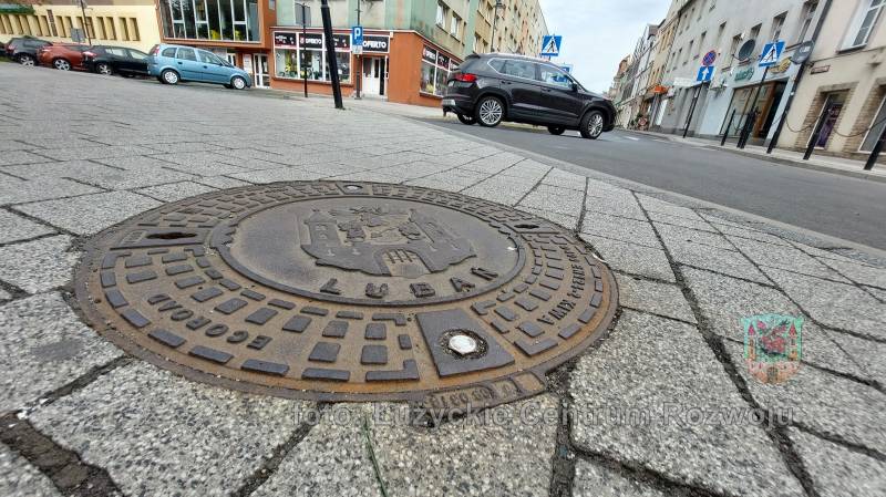 studzienka kanalizacyjna z herbem i napisem Lubań, w tle ulica